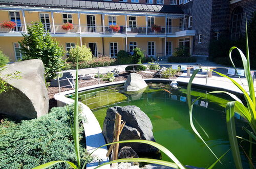 Teich mit grünem Wasser und großen Steinen vor einem gelben Gebäude mit mehreren Fenstern und Blumenkästen