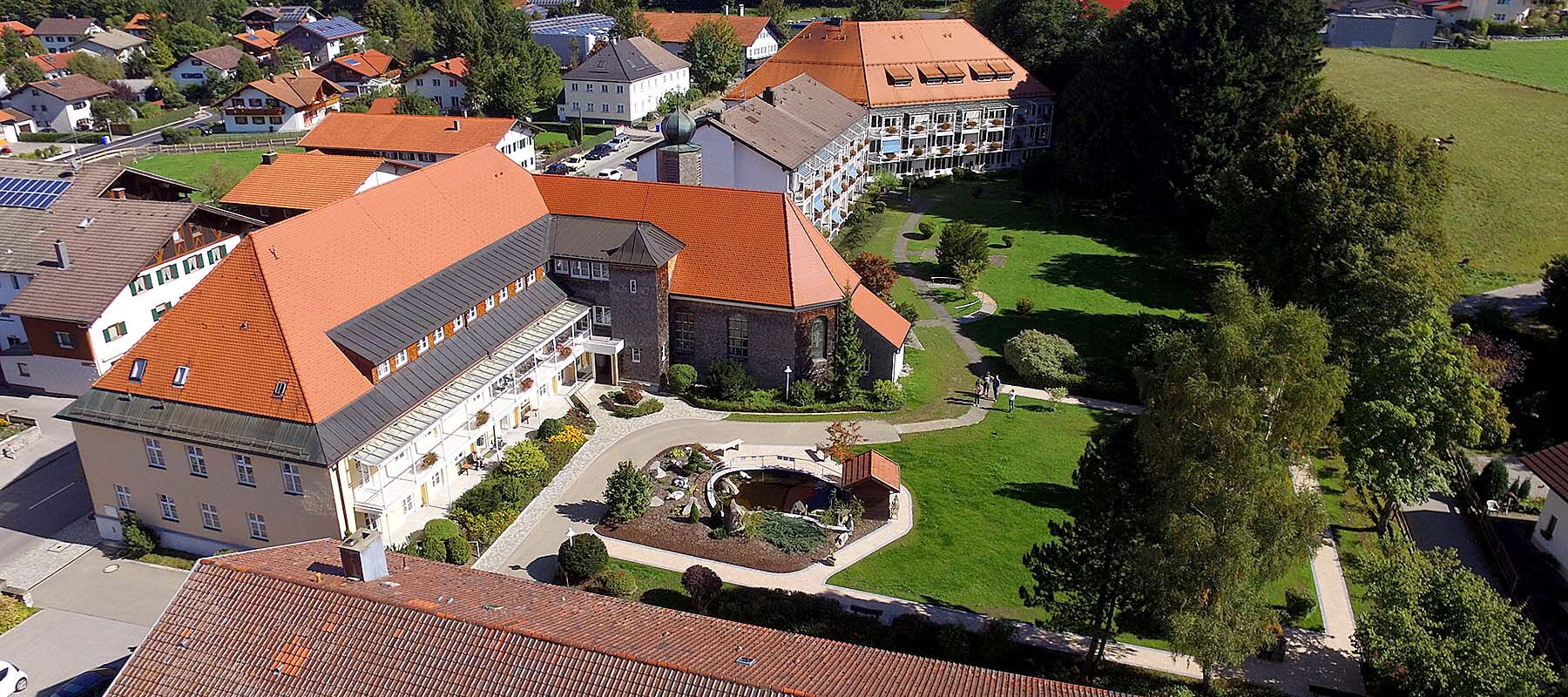 Luftaufnahme vom Pflegeheim mit roten Dächern, umgeben von Bäumen und Wiesen, im Hintergrund Berge