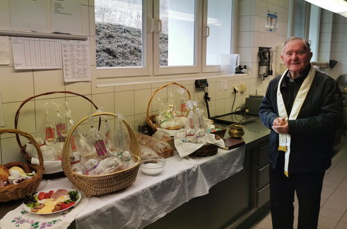 Person mit liturgischem Gewand steht neben einem Tisch mit mehreren gefüllten Weidenkörben und gedecktem Essen in einem Raum mit großen Fenstern