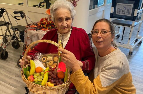 Ältere Person im Rollstuhl hält einen großen Korb mit Obst und Gebäck, neben ihr kniet eine jüngere Person in gelb-grauem Pullover