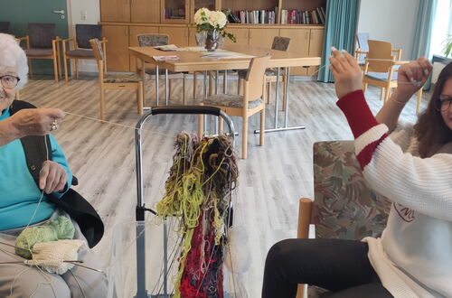 Zwei Personen sitzen sich in einem hellen Raum gegenüber und halten jeweils einen Faden in der Hand, zwischen ihnen hängt ein Knoten aus bunter Wolle.