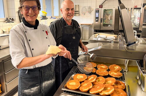 Zwei Personen in Kochkleidung stehen in einer professionellen Küche und wenden mit einem Sieb frittierte runde Teigstücke auf einem Backblech