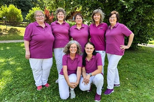 Gruppe von sieben Frauen in lila Poloshirts und weißen Hosen steht und kniet auf einer Wiese vor einem Holzzaun und Büschen