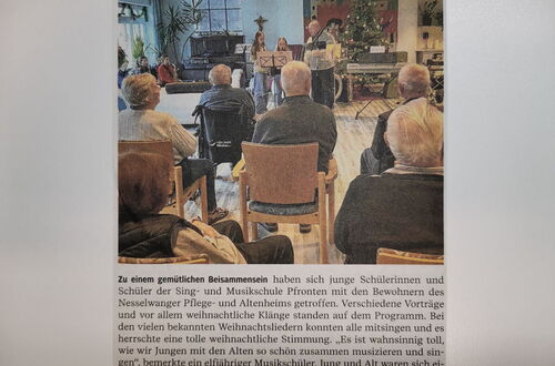 Mehrere ältere Menschen sitzen in einem Raum und hören einem kleinen Ensemble von jungen Musikerinnen und Musikern zu, die vor einem Weihnachtsbaum musizieren