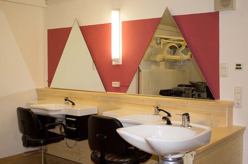 Friseursalon mit zwei weißen Waschbecken, schwarzen Friseurstühlen und dreieckigen Spiegeln an einer roten Wand