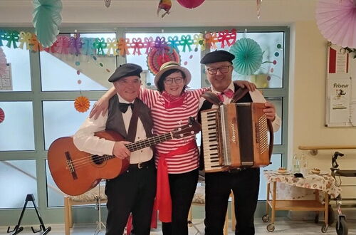 Drei Personen in Kostümen stehen eng umarmt in einem Raum mit bunten Papiergirlanden und -dekorationen an der Decke, zwei spielen Gitarre und Akkordeon, die mittlere trägt ein rot-weiß gestreiftes Hemd und eine Mütze