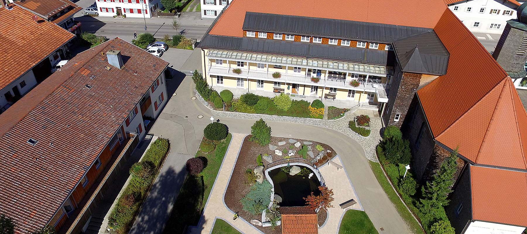 Luftaufnahme eines Gebäudekomplexes mit roten Dächern, einem Innenhof mit Teich und umlaufenden Wegen
