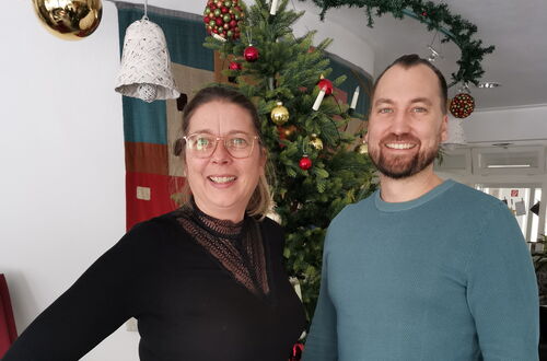 Zwei Personen stehen vor einem geschmückten Weihnachtsbaum in einem Raum mit Holzboden und roten Sitzmöbeln