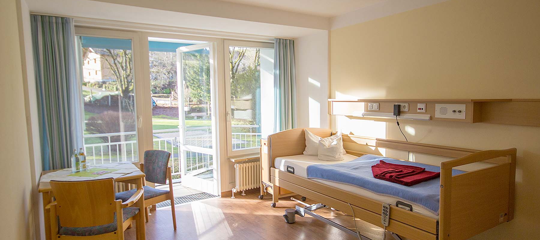 Ein helles Patientenzimmer mit einem verstellbaren Krankenhausbett, einem kleinen Tisch mit Stühlen und einer großen Fensterfront mit Blick ins Freie