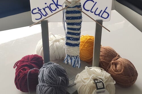 Stricknadeln mit einem kleinen, blau-weiß gestreiften Strickstück auf einem Ständer mit dem Schild Strick Club umgeben von mehreren Wollknäueln in verschiedenen Farben