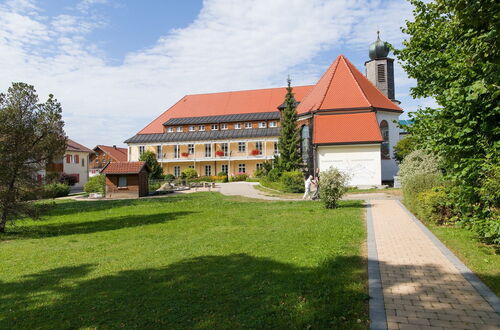 Großes Gebäude mit rotem Satteldach und kleinem Turm neben einem gepflasterten Weg auf einer grünen Wiese unter blauem Himmel