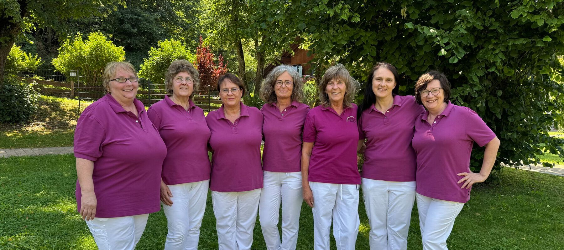 Sieben Frauen in lila Poloshirts und weißen Hosen stehen nebeneinander auf einer Wiese vor Bäumen