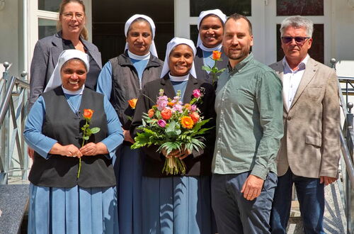 Gruppe von sechs Personen, darunter vier Frauen in blauen Kleidern und weißen Hauben. Schwester Amrutha hält einen Blumenstrauß, zwei Männer und eine Frau in Freizeitkleidung, alle stehen vor einem Gebäude mit Treppe