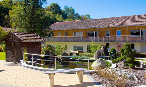 Garten mit Teich, kleiner Holzbrücke, Steinskulptur und zweistöckigem Gebäude im Hintergrund
