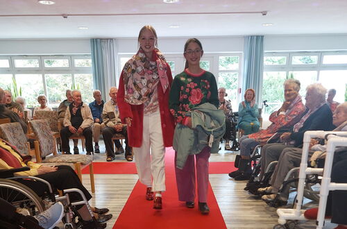 Zwei Frauen gehen auf einem roten Teppich in einem Raum mit sitzenden älteren Menschen, einige im Rollstuhl, die an den Seiten zuschauen