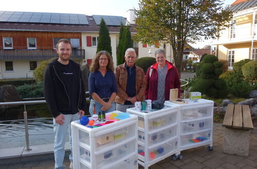 Vier Personen stehen hinter drei weißen Kunststoffkommoden mit verschiedenen Gegenständen darauf in einem Außenbereich mit Gebäuden und Bäumen im Hintergrund