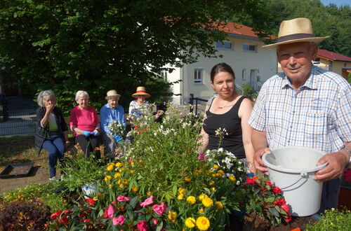Gruppe älterer Menschen sitzt auf einer Bank im Garten neben einem Beet mit bunten Blumen, im Vordergrund steht ein Mann mit Strohhut und Eimer, daneben eine Frau in schwarzem Top