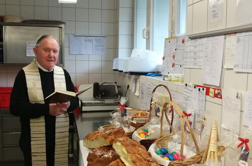 Pfarrer mit liturgischem Gewand steht in einer Küche vor einem Tisch mit Osterkörben gefüllt mit Eiern und Gebäck