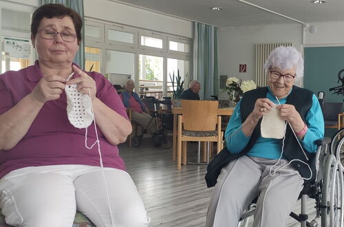 Zwei ältere Frauen sitzen in einem Gemeinschaftsraum und stricken jeweils ein weißes Stück Stoff