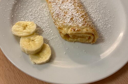 Auf weißem Teller ein gerollter Pfannkuchen mit Puderzucker bestreut und daneben drei Bananenscheiben auf hellem Holztisch