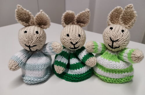 Drei gestrickte Hasenfiguren mit gestreiften Pullovern in Blau, Grün und Weiß auf weißer Oberfläche