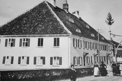 Großes historisches Gebäude mit Satteldach und mehreren Fenstern, davor stehen mehrere Personen in traditioneller Kleidung