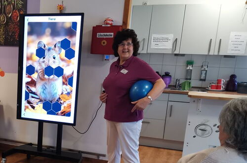 Frau mit lila Oberteil und weißer Hose hält blauen Ball vor Bildschirm mit Tierbild in einem Raum mit Holzfußboden und Küchenzeile, zwei ältere Personen sitzen im Vordergrund