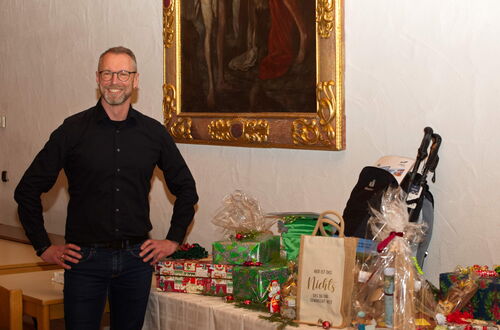 Mann in schwarzem Hemd steht mit Händen in der Hüfte vor einem Tisch mit Geschenken und Tragetaschen in einem Raum mit einem großen, goldgerahmten Gemälde an der Wand