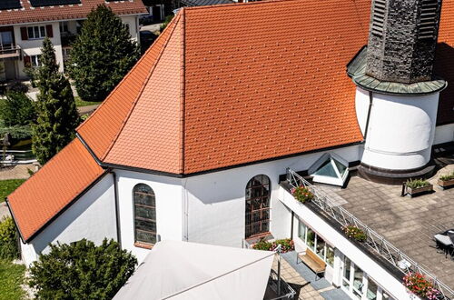 Luftaufnahme einer Kirche mit rotem Satteldach, rundem Turm und angrenzendem Gebäude mit Terrasse und Sonnensegel