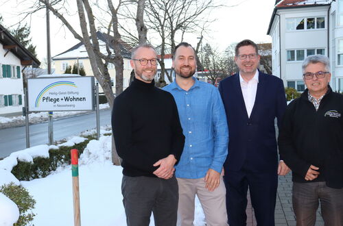 Vier Männer stehen vor einem Schild der Heilig-Geist-Stiftung Pflege + Wohnen in Nesselwang im Winter mit Schnee auf dem Boden