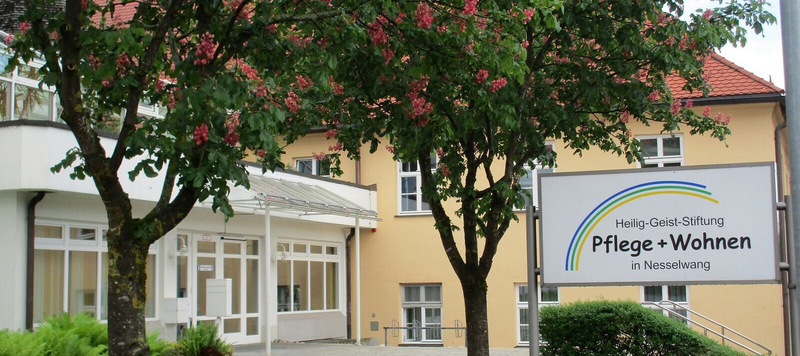 Eingangsbereich eines Gebäudes mit zwei blühenden Bäumen und einem Schild mit der Aufschrift 'Heilig-Geist-Stiftung Pflege + Wohnen in Nesselwang'