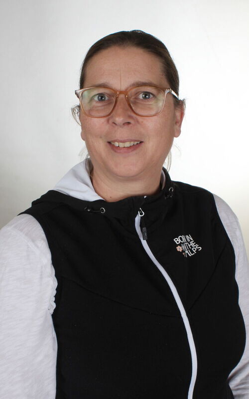 Frau mit Brille trägt eine schwarze Weste mit weißem Reißverschluss und weißem Text auf der linken Brust über einem hellen Kapuzenpullover