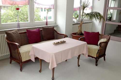 Sitzgruppe mit zwei Korbsesseln und einem Korbsofa um einen Tisch mit hellrosa Tischdecke in einem hellen Raum mit großen Fenstern und Pflanzen