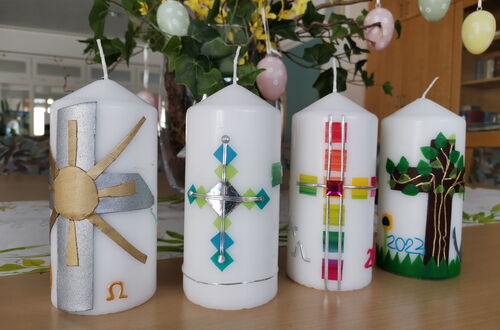 Vier weiße Kerzen mit verschiedenen bunten Symbolen wie Sonne, Kreuz, Regenbogenkreuz und Baum stehen auf einem Holztisch