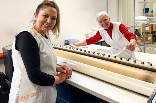 Zwei Frauen in weißen Schürzen bedienen eine lange Maschine mit mehreren Knöpfen in einem Arbeitsraum