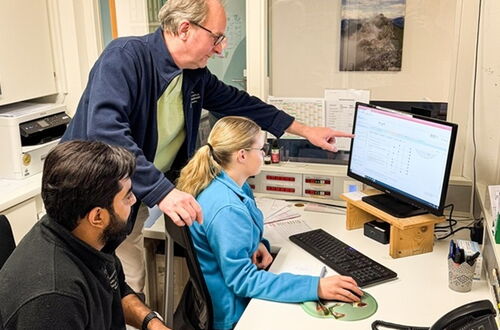 Drei Personen sitzen an einem Schreibtisch, eine Person zeigt auf einen Computerbildschirm mit Tabellen und Diagrammen