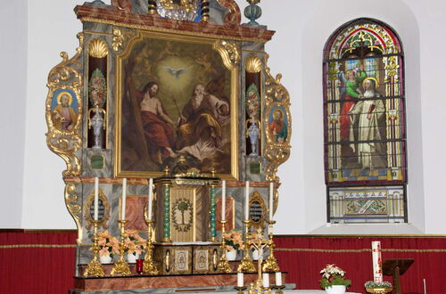 Barocker Altar mit Gemälde der Heiligen Dreifaltigkeit, flankiert von vergoldeten Verzierungen und Kerzen, daneben ein buntes Kirchenfenster mit einer Heiligenfigur