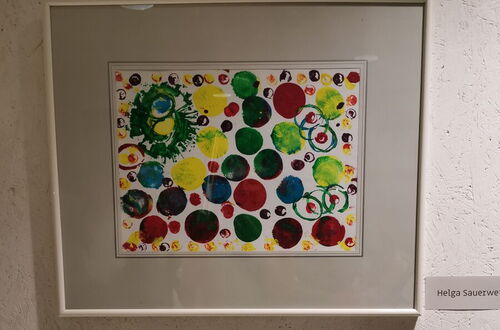 Zwei gerahmte Kunstwerke an einer Wand, oben ein farbiges Bild mit vielen bunten Kreisen, unten zwei schwarz-weiße Porträtzeichnungen mit unkenntlich gemachten Gesichtern