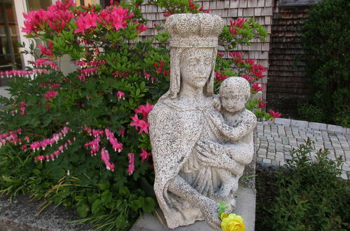 Steinerne Statue einer gekrönten Frau mit Kind auf dem Arm vor blühendem Rhododendron und Backsteinmauer
