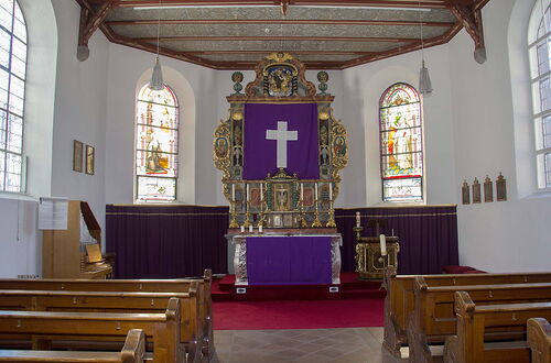 Innenraum einer Kirche mit Altar, der mit einem lila Tuch mit weißem Kreuz bedeckt ist, umgeben von bunten Glasfenstern und Holzbänken