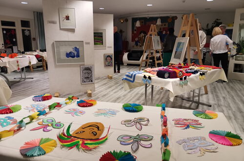 Ausstellung mit bunten Bastelarbeiten auf Tischen in einem hellen Raum mit mehreren Personen im Hintergrund
