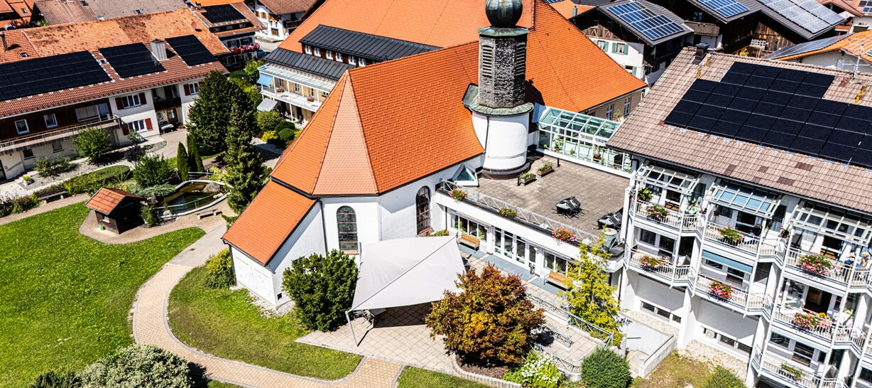 Luftaufnahme eines Gebäudekomplexes mit Kirche, roten Dächern und Solarpaneelen auf einem der Dächer