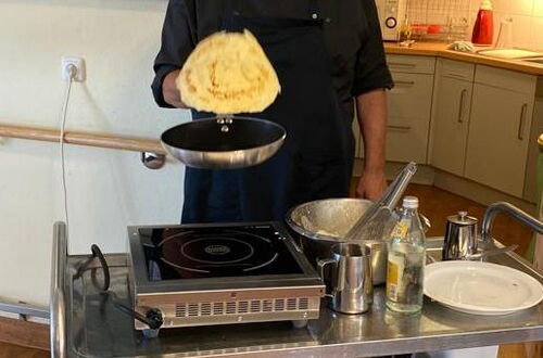 Mann in schwarzer Kochjacke hält Pfanne mit fliegendem Pfannkuchen vor Küchenschränken und Tafel mit Zeichnungen