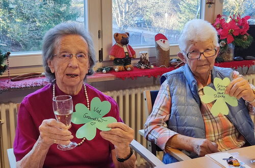 Zwei ältere Frauen sitzen an einem Tisch, eine hält ein Glas und ein grünes Kleeblatt mit der Aufschrift 'Viel Gesundheit', die andere ein grünes Kleeblatt mit der Aufschrift 'Gesundheit und Stärke'