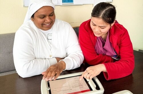 Zwei Frauen sitzen an einem Tisch und betrachten gemeinsam ein Tablet, eine trägt eine weiße Arbeitskleidung, die andere eine rote Jacke