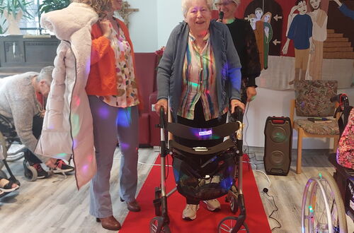 Zwei Frauen stehen auf einem roten Teppich, eine davon mit Rollator, im Hintergrund weitere Personen und eine Wand mit einem Wandteppich