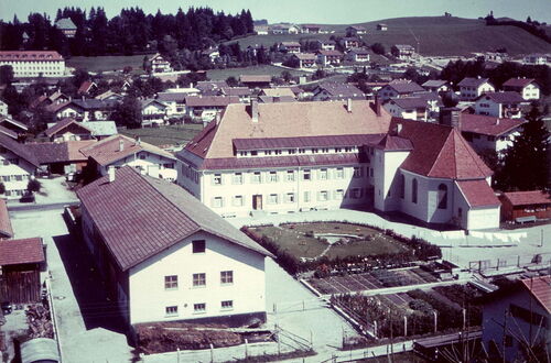 Blick auf das Pflegeheim mit mehreren Gebäuden, darunter ein großes Haus mit rotem Dach und eine Kirche mit spitzem Turm, umgeben von Hügeln und Wald