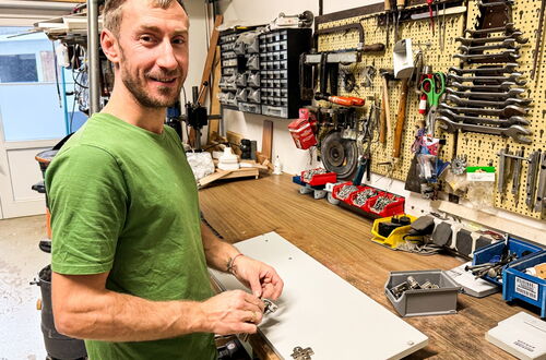 Mann in grünem T-Shirt steht an Werkbank mit Werkzeugwand und hält ein kleines Metallteil in der Hand