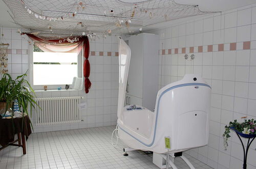 Weiß gekachelter Raum mit einem großen weißen Badewannenähnlichen Gerät auf Rollen, einem Fenster mit rotem Vorhang und mehreren Pflanzen auf Tischen